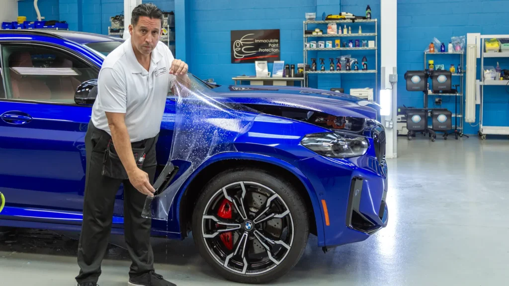 Bill Fetter demonstrating a hands-on SunTek Reaction PPF installation on a San Marino Blue BMW X4M Competition. This shot highlights the real-world craftsmanship and clean, professional studio environment required for a high-tier, 12-year warranty paint protection finish.