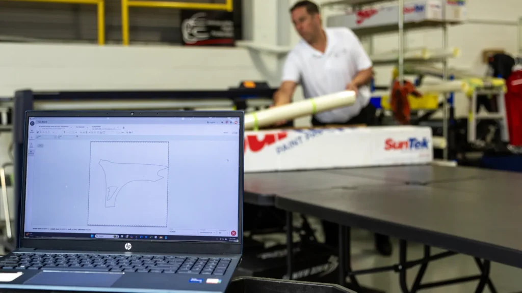 Bill Fetter preparing a roll of SunTek Reaction PPF for the Graphtec plotter. The laptop in the foreground displays the engineered fender pattern for a BMW X4M Competition, showing the start of the precision cutting process that eliminates the need for hand-trimming on the vehicle's paint.