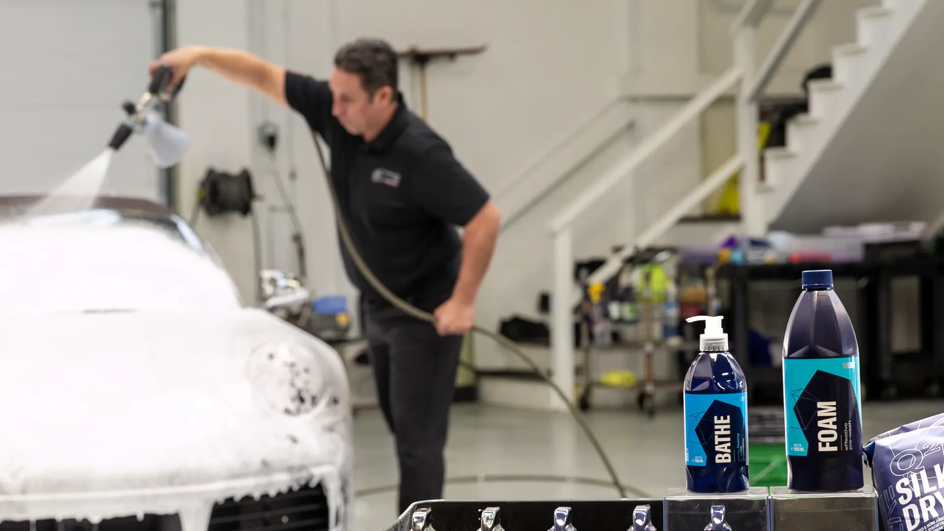 When teaching the best way to hand wash car exteriors safely, Bill Fetter, founder of Immaculate Paint Protection, trusts premium detailing products. This close-up highlights a Gyeon foam bottle in the foreground, while Bill Fetter uses a foam cannon to apply a thick pre-soak to a 2020 Porsche 911 Convertible in the background. Using high-quality foam before washing a car by hand is a critical step experts use to lift dirt and prevent paint damage.