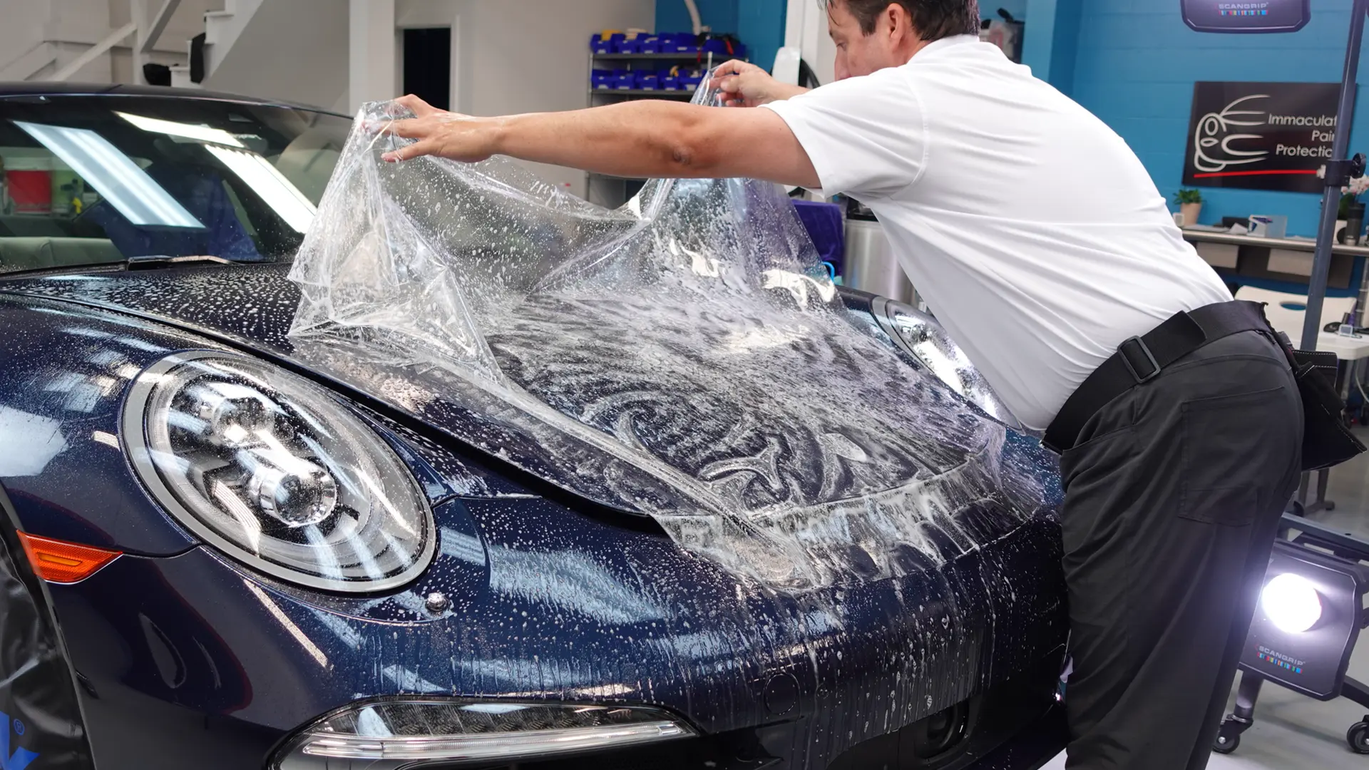Bill Fetter of Immaculate Paint Protection carefully aligning a clear sheet of Paint Protection Film (PPF) onto the hood of a 2014 Porsche 911 inside the shop.