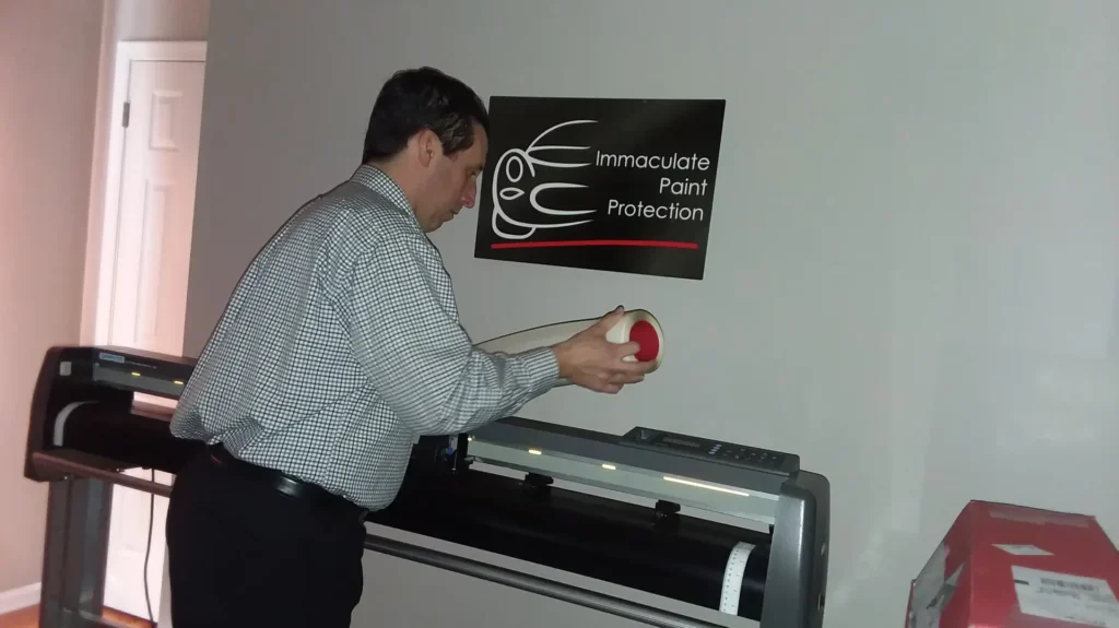 A throwback photo from 2018 showing Bill Fetter loading paint protection film into his first plotter in his home office, demonstrating the early days of Immaculate Paint Protection.