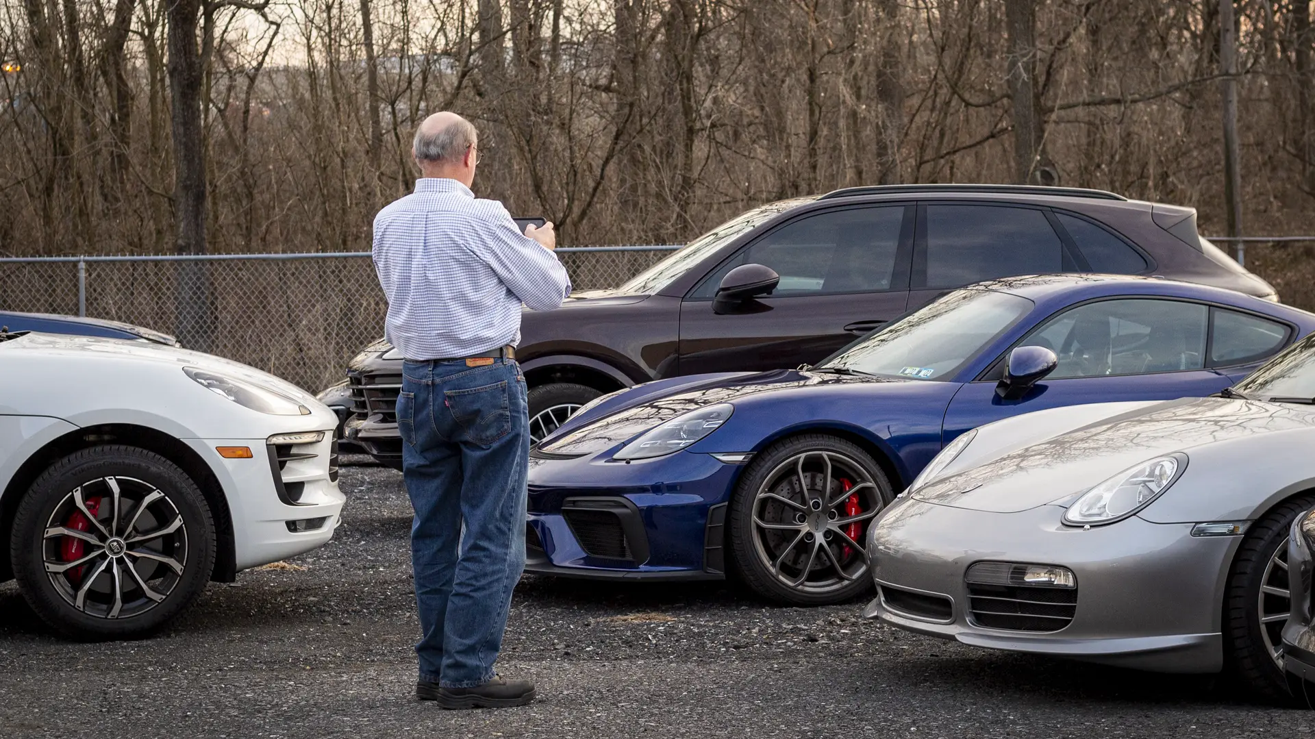 A Porsche Club member taking a photo of a Porsche 718 Cayman in the Immaculate Paint Protection parking lot, surrounded by a Porsche 911, Macan, and Cayenne during a club meeting.