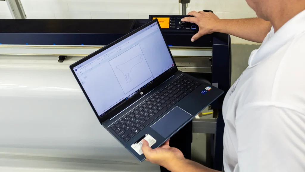 Bill Fetter of Immaculate Paint Protection standing at a professional Graphtech CNC plotter, reviewing a BMW X4M Competition fender PPF pattern on a laptop before cutting the film.