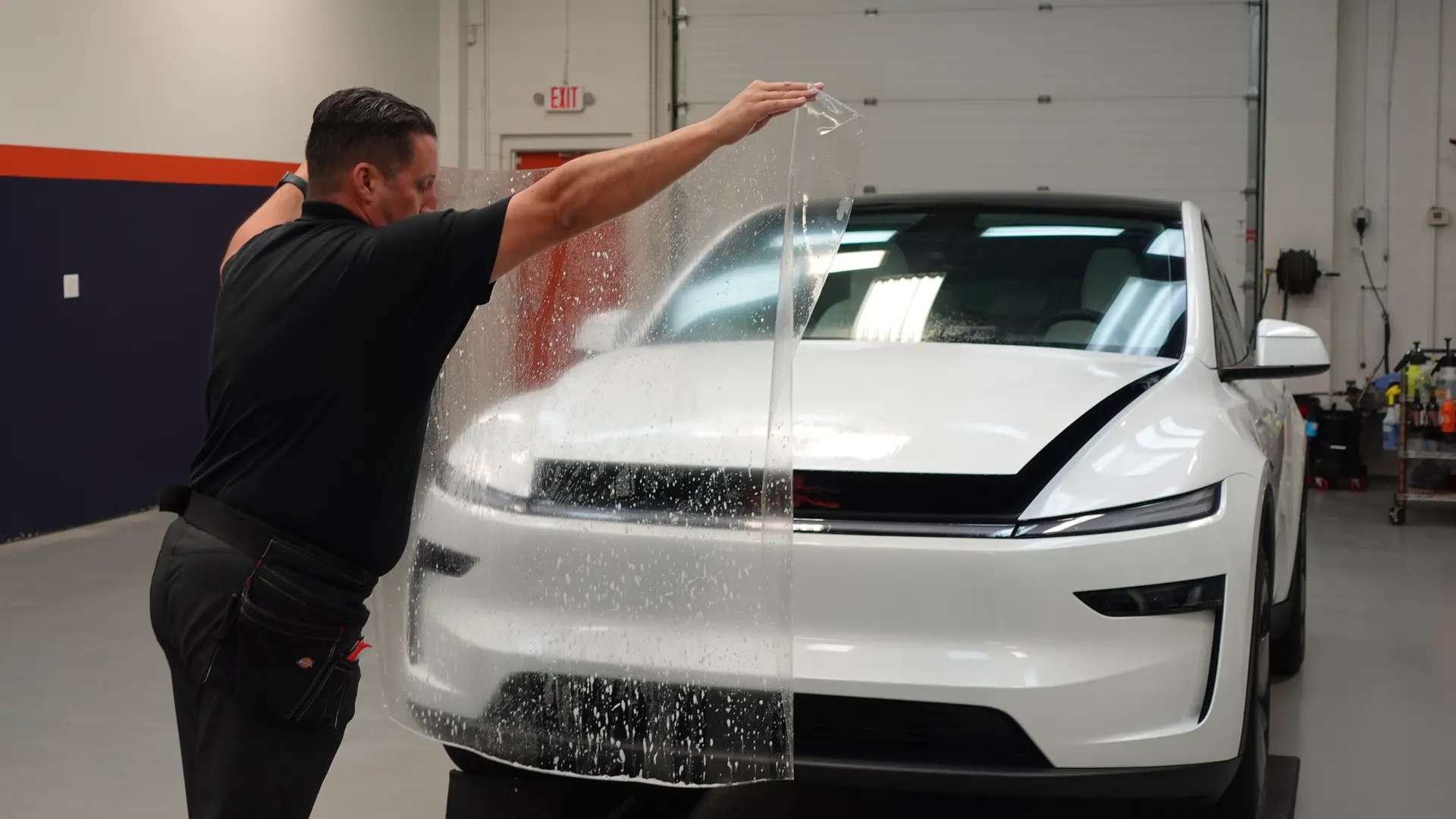 Bill Fetter of Immaculate Paint Protection carefully aligning a clear sheet of Paint Protection Film (PPF) onto the hood of a 2026 Pearl White Multi-Coat Tesla inside the shop.